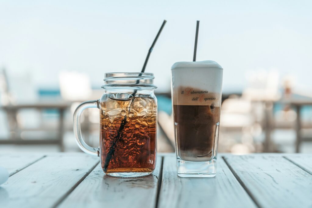 Buzlu çay mason jar ve köpüklü soğuk kahve ahşap masada deniz manzarası